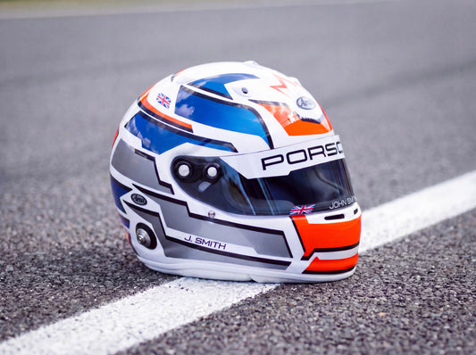 A white, blue, and orange racing helmet with a clear visor resting on an asphalt racetrack next to a white painted line. The helmet features 'PORSCHE' and 'J.